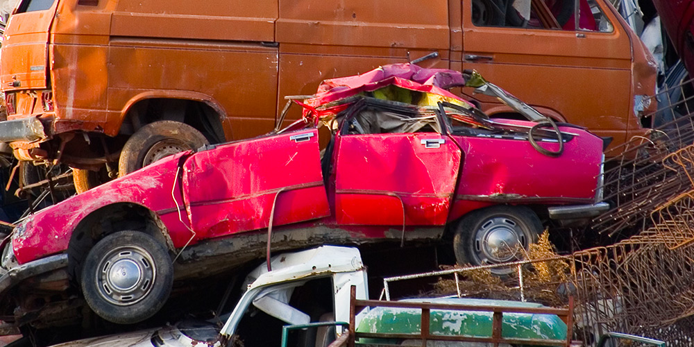 Auto wrecking yard with orange van, crushed red convertible and white pickup truck between scrap metal