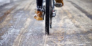 A close up of the rear of a bicycle on the road. There is slushy snow on the ground that is being kicked up by the rear tire. The rider is wearing brown boots and black pants.