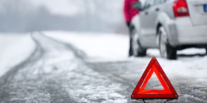 A red and neon orange hazard triangle is on a snowy road. There is a silver car in the background with a person standing in front of the car.