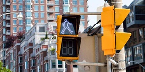 Two square black and yellow signal lights attached to a grey street post with the pedestrian walk sign illuminated in white at the top. There is a second set of yellow and black signal lights facing to the right on the same pole. Behind is a row of red and white buildings and a high rise apartment. 