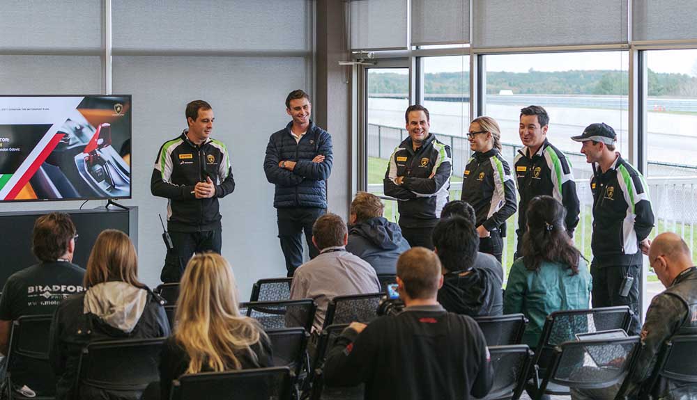 Conference room with Lamborghini team in race car suits at front led by Paolo Biglieri