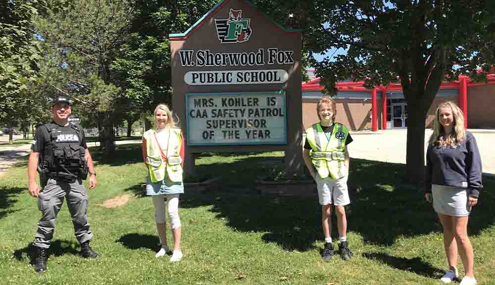 Three students and a police officer flank a sign in front of a school. The school sign, W. Sherfood Fox Public School, reads: Mrs. Kohler is CAA Safety Patrol Supervisor of the Year.