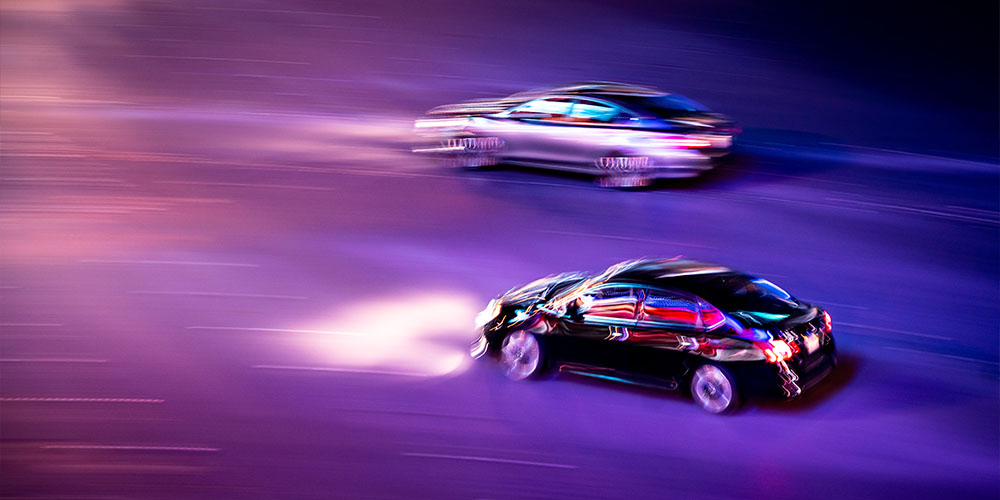 Side profiles of two blurred cars against a purple background. There is a silver car in the background, and a black sedan parallel to it with its headlights on.