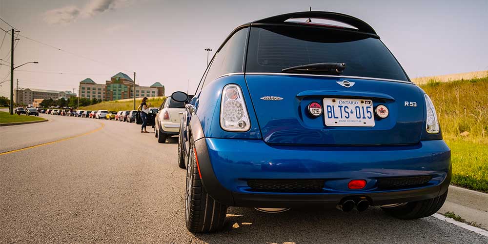 The back of a bright blue Mini Cooper hatchback vehicle with an Ontario licence plate sits parked on the side of a road next to a field. There is a row of parked cars along the curved street, which leads to a building complex in the distance. A woman is seen opening a car door ahead.