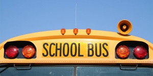 The upper lights of a school bus. The school bus is yellow with the words school bus written in black capital letters in the centre front of the bus with two circular red lights on either side. There are three amber lights on the top centre of the bus. A horn is fixed to the top right on the roof. You can see the top of the front windshield at the bottom and a blue background behind. 