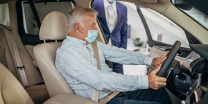 A man with silver hair wearing a light blue button up shirt and jeans, wearing a mask, is sitting in a car. He has a seat belt on and is looking at the steering wheel. There is another man standing behind him with an open car door wearing a white button up shirt and a bright blue suit.