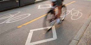A close up of a road, which is marked with a white outline of a diamond and a white bicycle. There is also a yellow road marking next to the one white diamond, with a white bicycle outline on the other side of it. A blurred out bicycle and pair of feet can be seen riding in one of the lanes.
