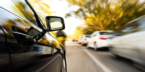 On the left, a profile of the passenger door and mirror of a black car, driving on a street past several white vehicles on the right, which are blurry to show a fast rate of speed. Blurry trees can be seen in the background.
