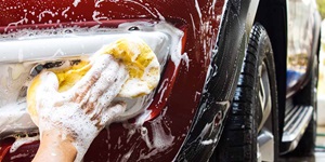 A close up of a hand that has lots of soap on it. The hand is holding up a soapy yellow sponge to the front lights of a red car. Other parts of the car have soap on it as well.