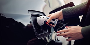 Close up of a woman’s hands that have red nail polish. She is wearing a dark green shirt. Her one hand is holding the handle of a white and black charger and she’s plugging it into the side of a black car.