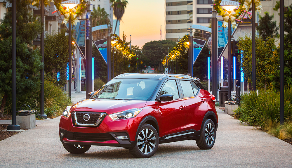 Cherry red Nissan Kicks crossover showcased in front of urban backdrop with high rises on tree-lined street at sunset
