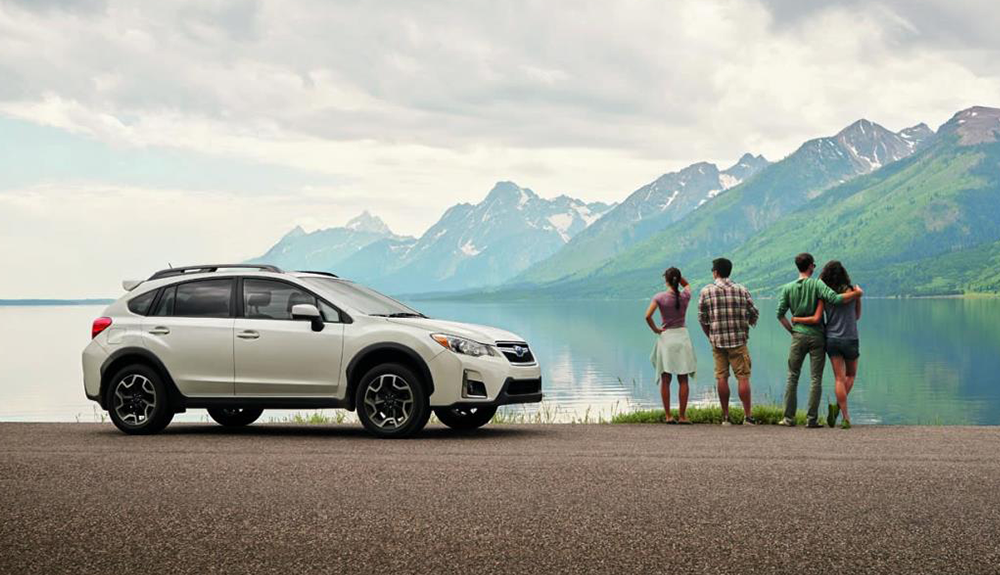 Two couples, one with arms around each other, gazing over still lake at snowy mountaintops beside a light silver Subaru Crosstek