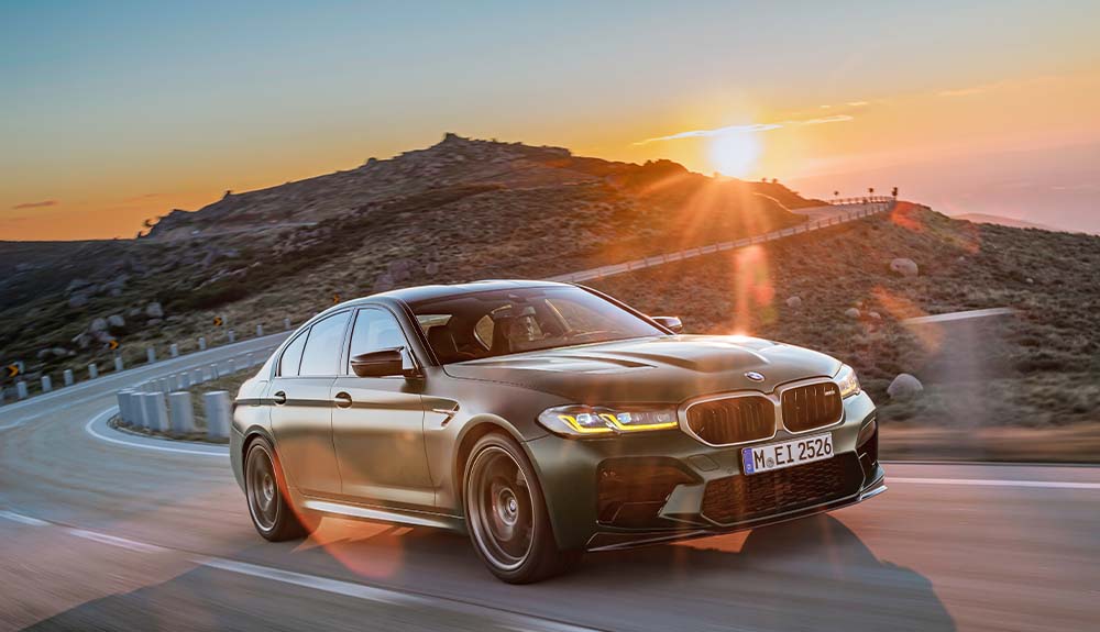 A matte green BMW M3 is shown driving on a two lane highway with hills in the background
