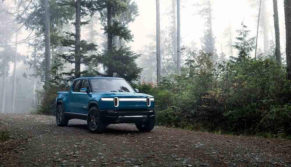 A blue 2022 Rivian R1T pickup truck is shown on a dirt road in a wooded area