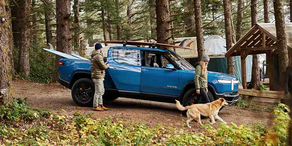A couple and their dog are shown unloading surfboards from a blue 2022 Rivian R1T pickup truck 