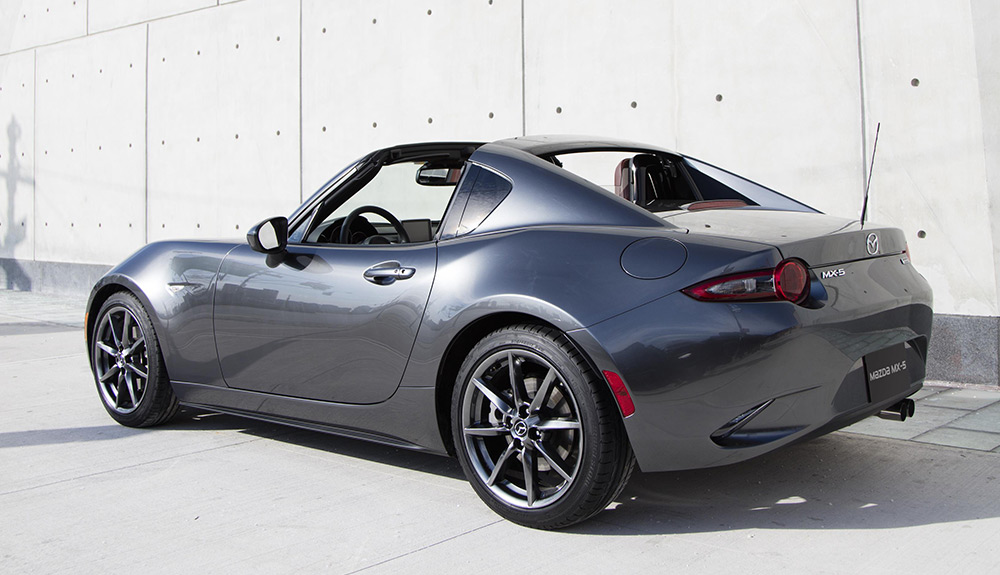 Back and side shot of a dark grey Mazda MX-5 RF parked against an industrial wall