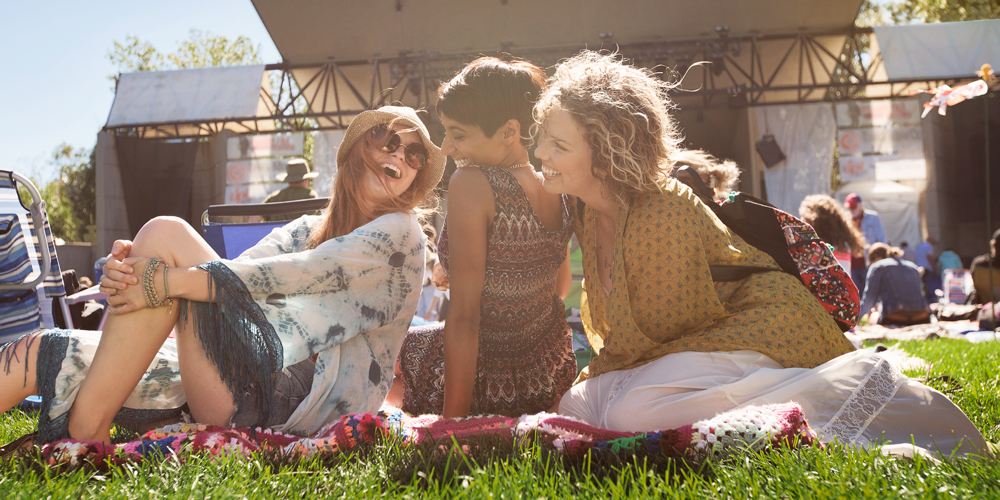 Three woman laugh together while sitting on blankets in the grass at an outdoor concert in Calgary, Alberta