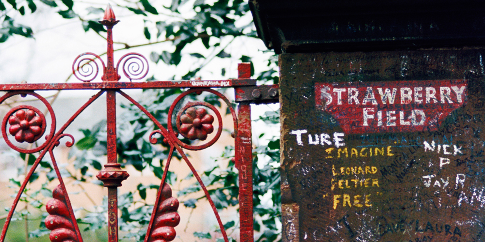 A painted red metal sculpture stands in front of a sign that reads Strawberry Field in Liverpool, England