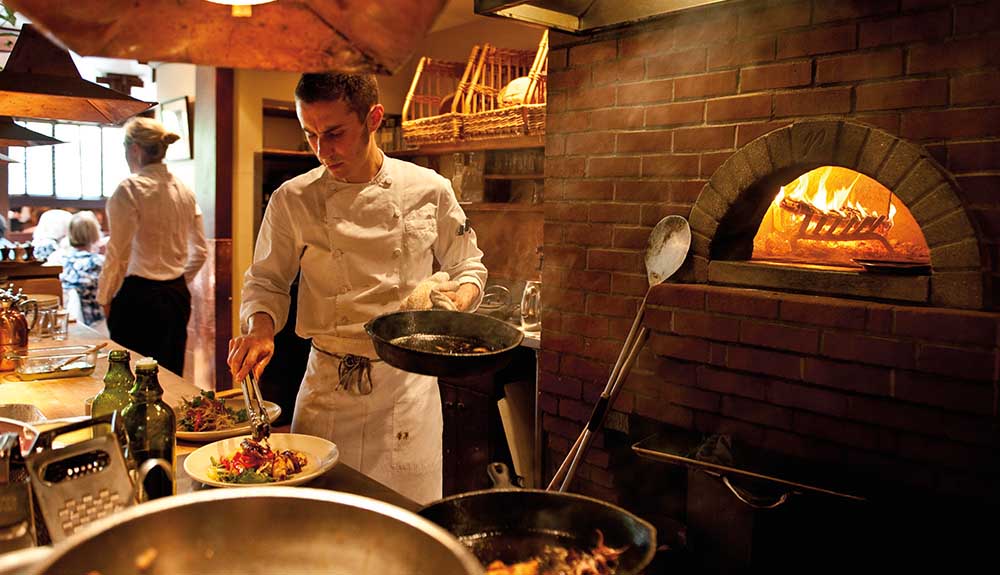 A man in chef's whites spoons sauce from a cast iron pan onto a elegantly plated dish, a wood pizza oven blazing behind him