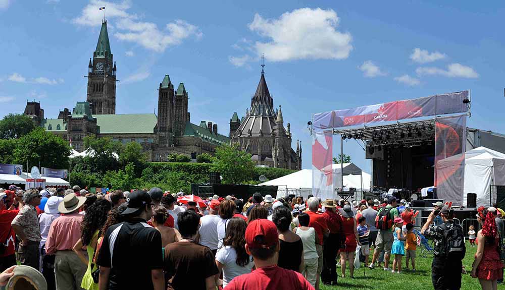 A crowd dressed in summer attire gather in front of a stage near parliament in Ottawa, Ontario