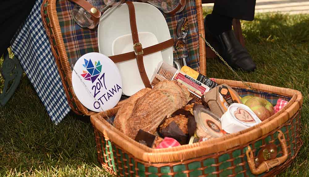 A picnic basket sits open displaying artisan products such as bread, cheese, apples, crackers and muffins, plates strapped in the top of the basket and a large button that reads 2017 Ottawa