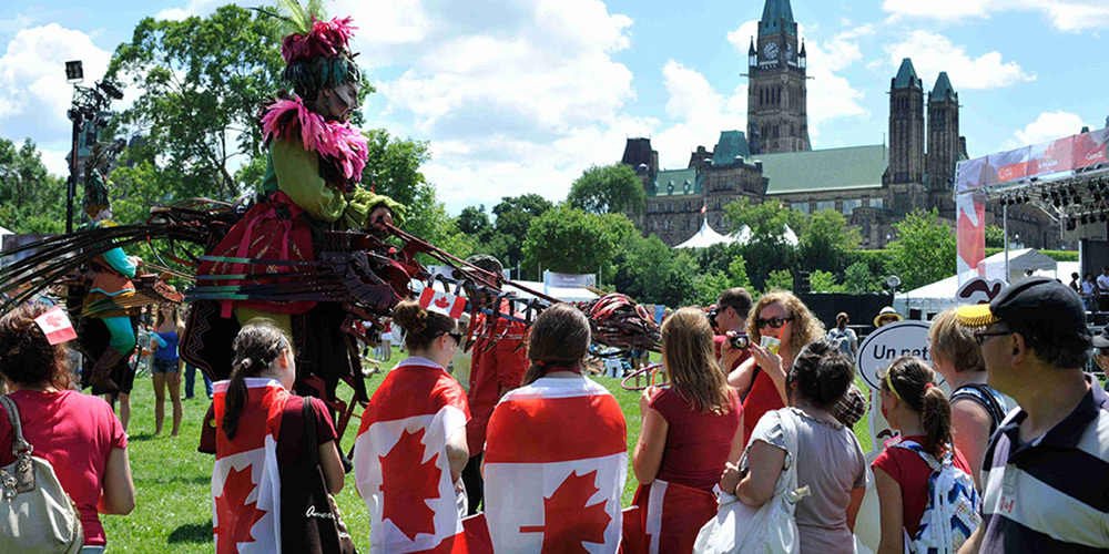 A crowd, many wearing Canadian flags or red shirts, gathers around a performer on stilts at an outdoor Canada Day celebration in Ottawa 