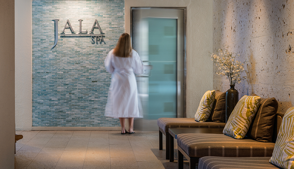 A woman in a white robe enters a glass door at the Jala Spa