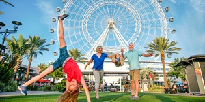 A young girl does a cartwheel in the grass in front of a man and woman swinging a young boy in ther arms, a large white ferris wheel behind them