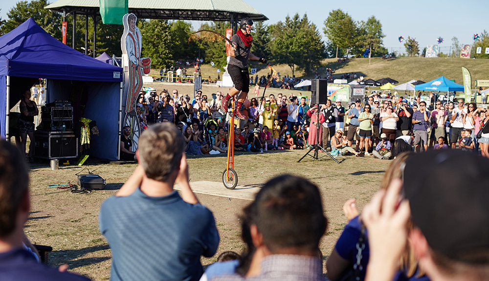 Man on a unicycle as the crowd captures the act with cameras and phones in the daytime at Busker's Fest