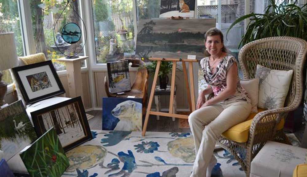 Woman sits on the edge of a whicker chair next to an empty easel, surrounded by art