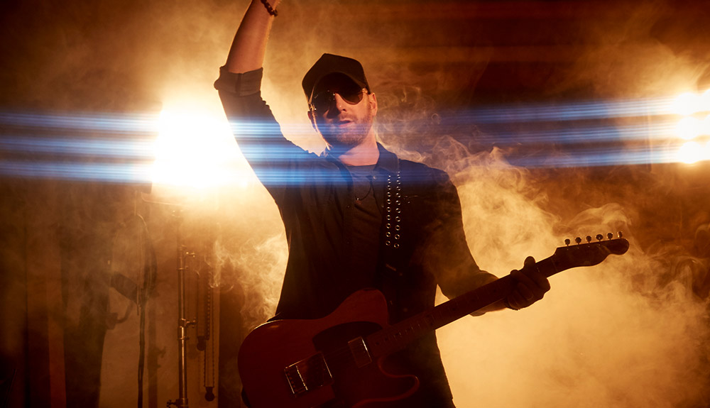 Country singer holding a guitar with sunglasses and a baseball cap holds his fist up as a smoke machine generates smoke behind him on stage