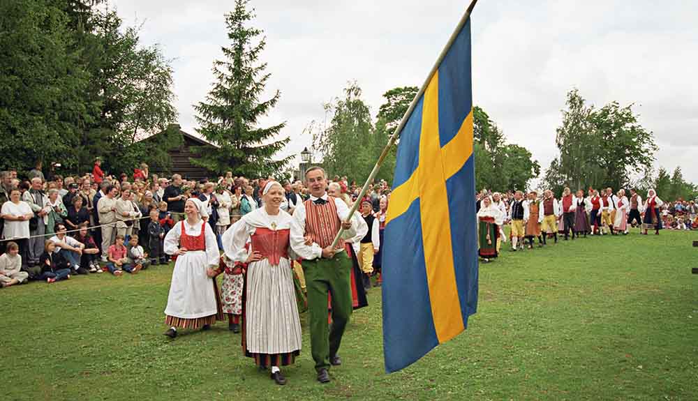 Parade of men and women dressed in traditional outfits, front two hold Sweden's flag as they celebrate Midsummer's Eve