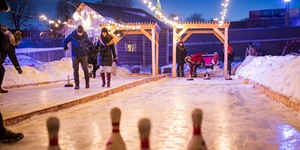 Two lanes of outdoor ice curling are seen illuminated with twinkle lights, a couple playing on one and a family on the second in Montreal