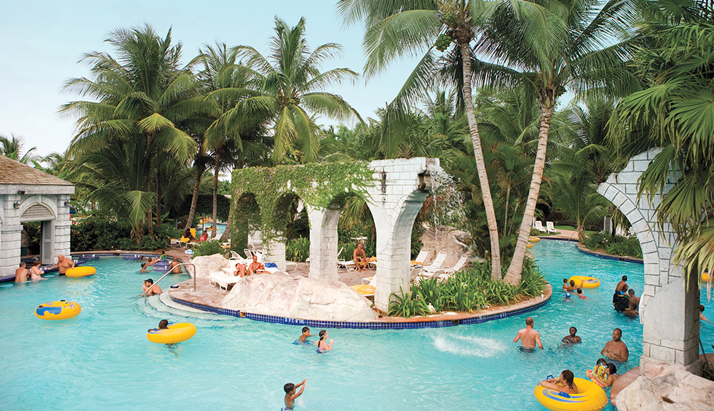A luxurious hotel pool in Jamaica with a small sandy island in the centre, happy visitors splashing and frolicking in the waters