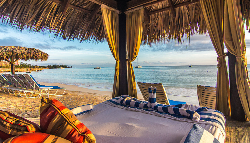 A beachfront tent shelters a day bed with pillows looks out at the calm beach