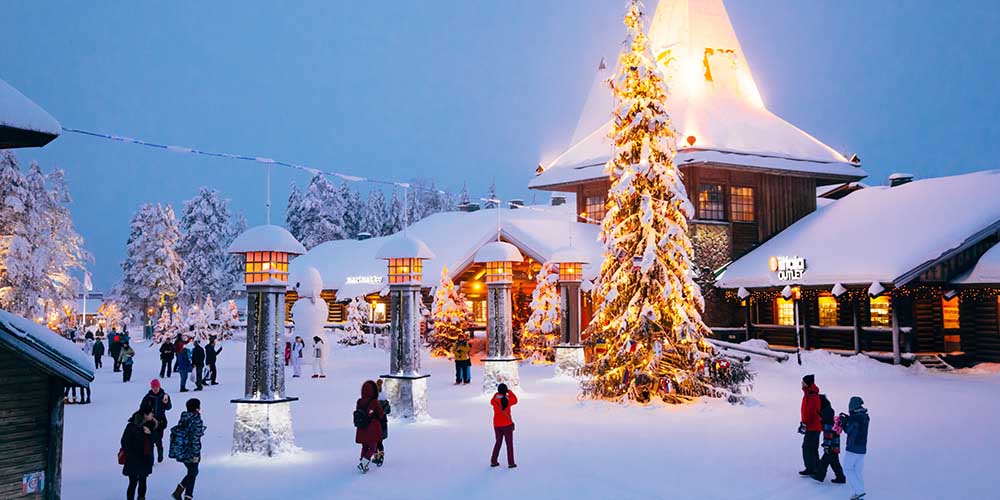 Tall outdoor Christmas tree stands outside of a row of lights and buildings covered in snow, with people around