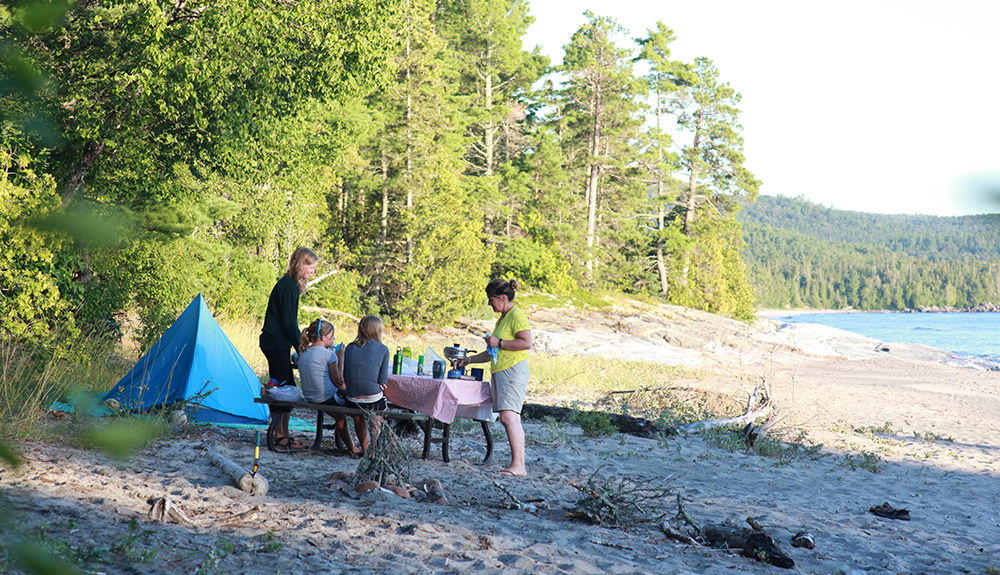 Two kids sit at a picnic table while two grown ups stand by serving eats next to a tiny blue tent on Agawa Beach