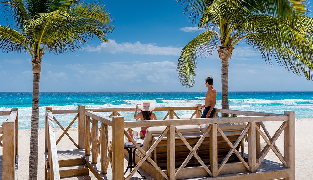 Woman in straw hat and man in bathing suit look out onto the beach and ocean from a resort patio with an outdoor bed