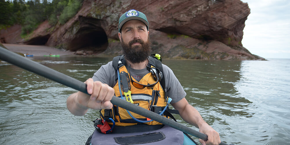 Bearded man wearing a baseball cap kayaks in a life jacket in the Bay of Fundy