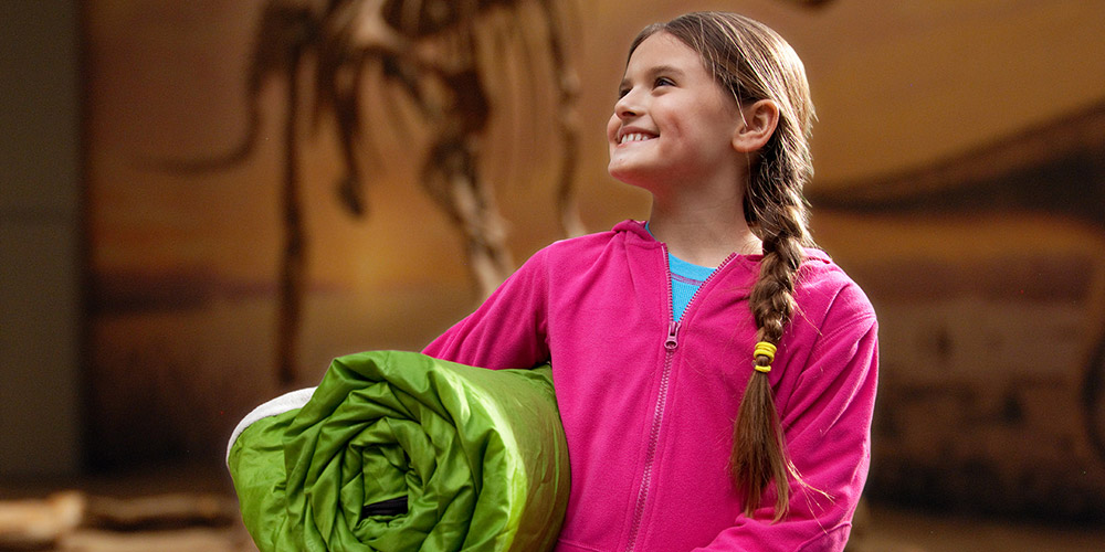Smiling little girl in a pink zip-up holding a rolled up green sleeping bag