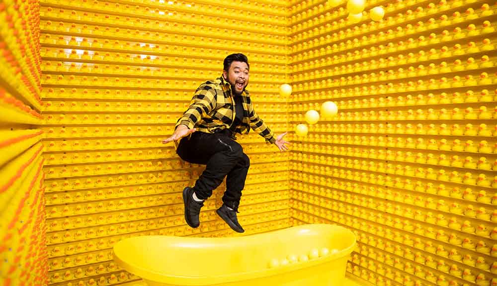 Yellow room lined with rubber duckies and a yellow bathtub filled with balls as a man in a yellow plaid jacket jumps into it