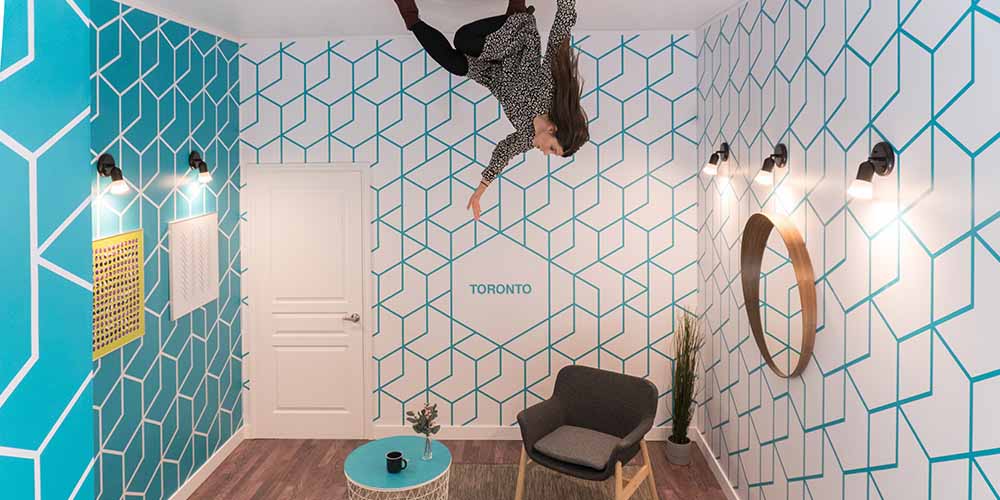 Woman enjoys herself at the Museum of Illusions in a photo where she appears to be standing on the ceiling with a table and chair on the ground
