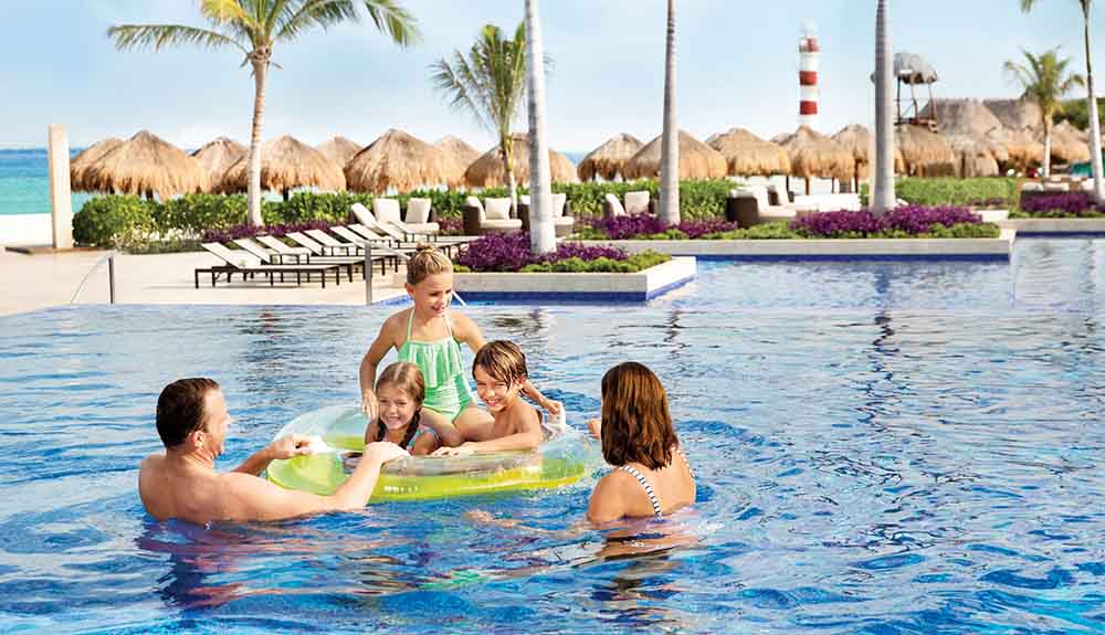 Three kids ride a floatie in a pool with a man helping and a woman watching