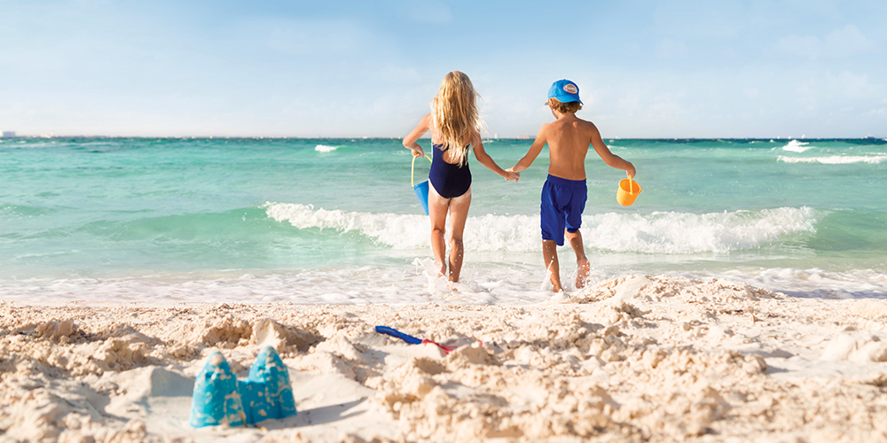 A little girl and little boy in swimwear carrying pails into the green-blue ocean on the beach