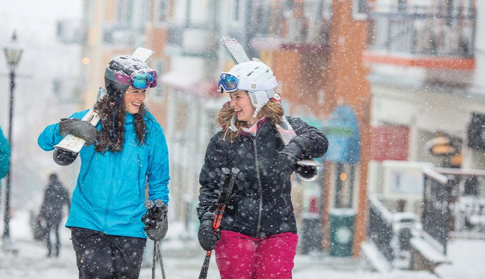 Two women carrying skis past shops in resort village at Mont Tremblant in Quebec Canada