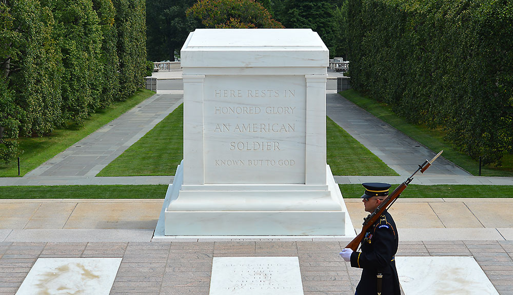 A soldier marches in front of a white monument that reads Here Rests In Honored Glory An American Soldier But Known to God