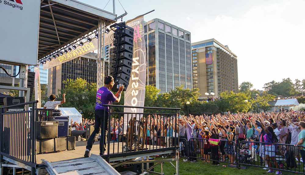 A crowd cheers as a DJ is seen playing on stage, a person filming to the side of them