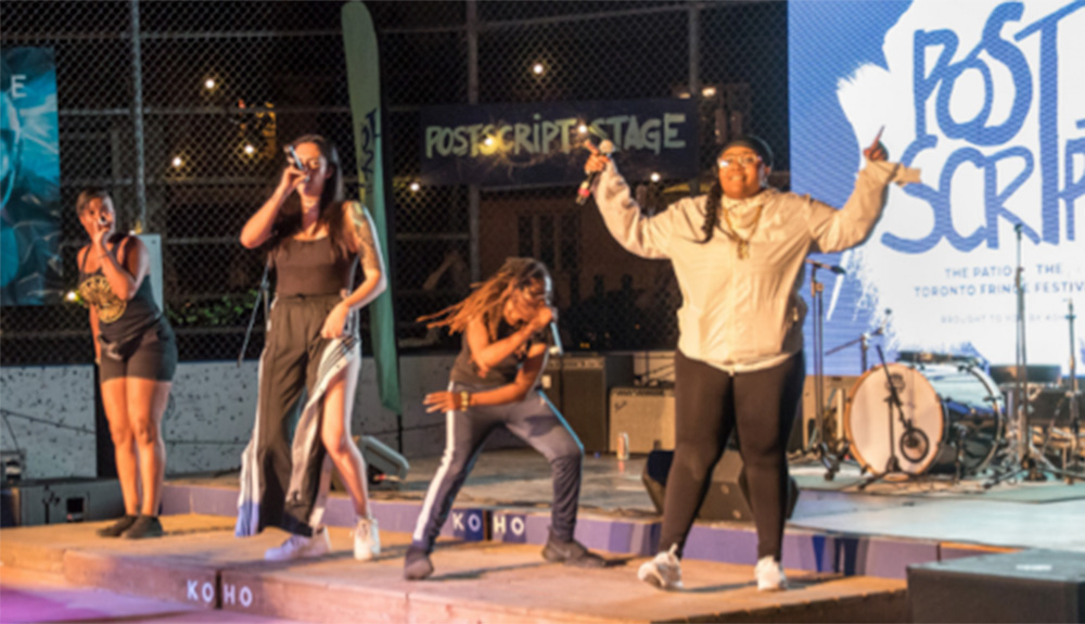 A performance on the PostScript Stage at the Toronto Fringe Festival featuring four women with microphones singing and dancing