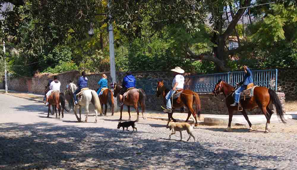People riding horses with dogs trailing along cobblestone streets in a small Mexican town