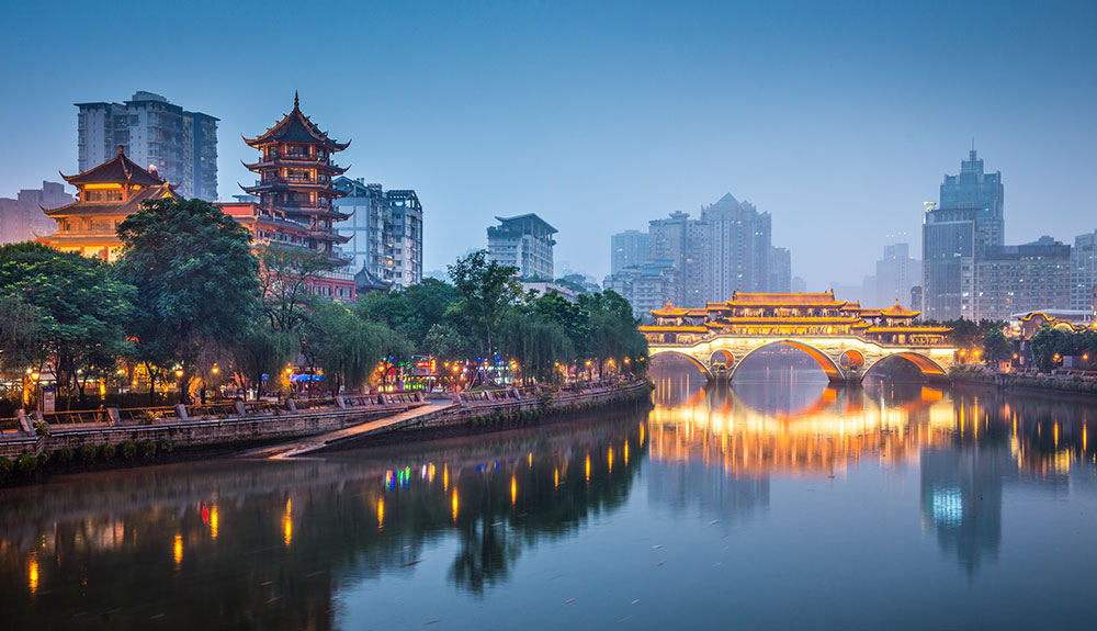 Pagodas behind trees along the coast as a bridge lit by pretty lights illuminates the water beneat it in Chengdu, China